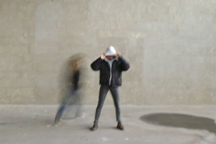 Fotografie einer Person, die vor einer gräulichen Wand steht und der verschwommene Umriss einer Person die um sie herum rennt.